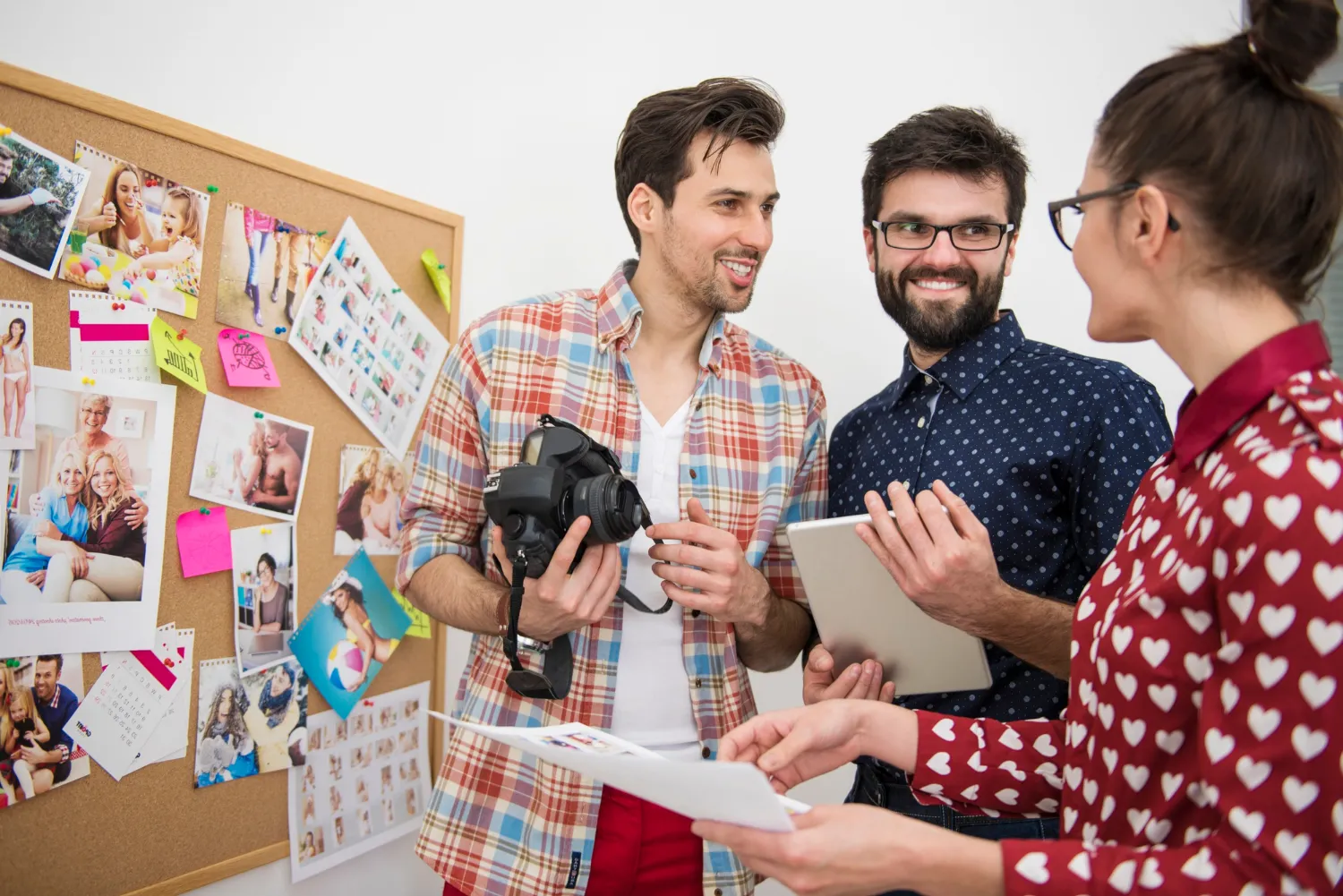 Grupo de tres personas colaborando en un proyecto fotográfico, sosteniendo una cámara y una tablet, mientras revisan imágenes en un tablero de corcho con fotos y notas.